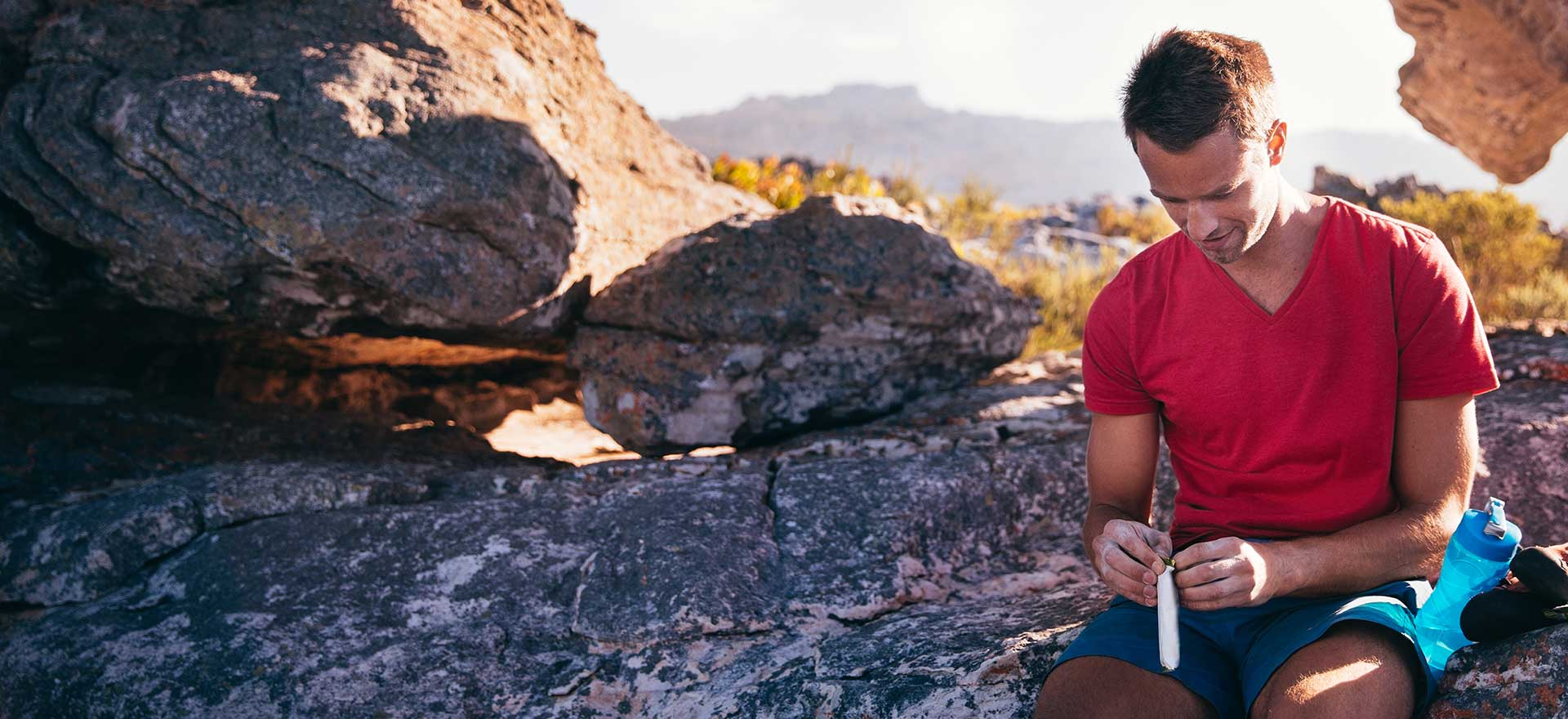 Man sitting on a rock while hiking, about to open a snack bar.