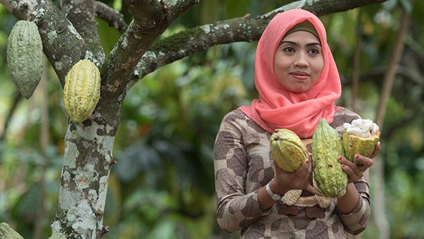 Indonesian woman smiling while standing next to a tree and holding fruits that have been picked from the tree.