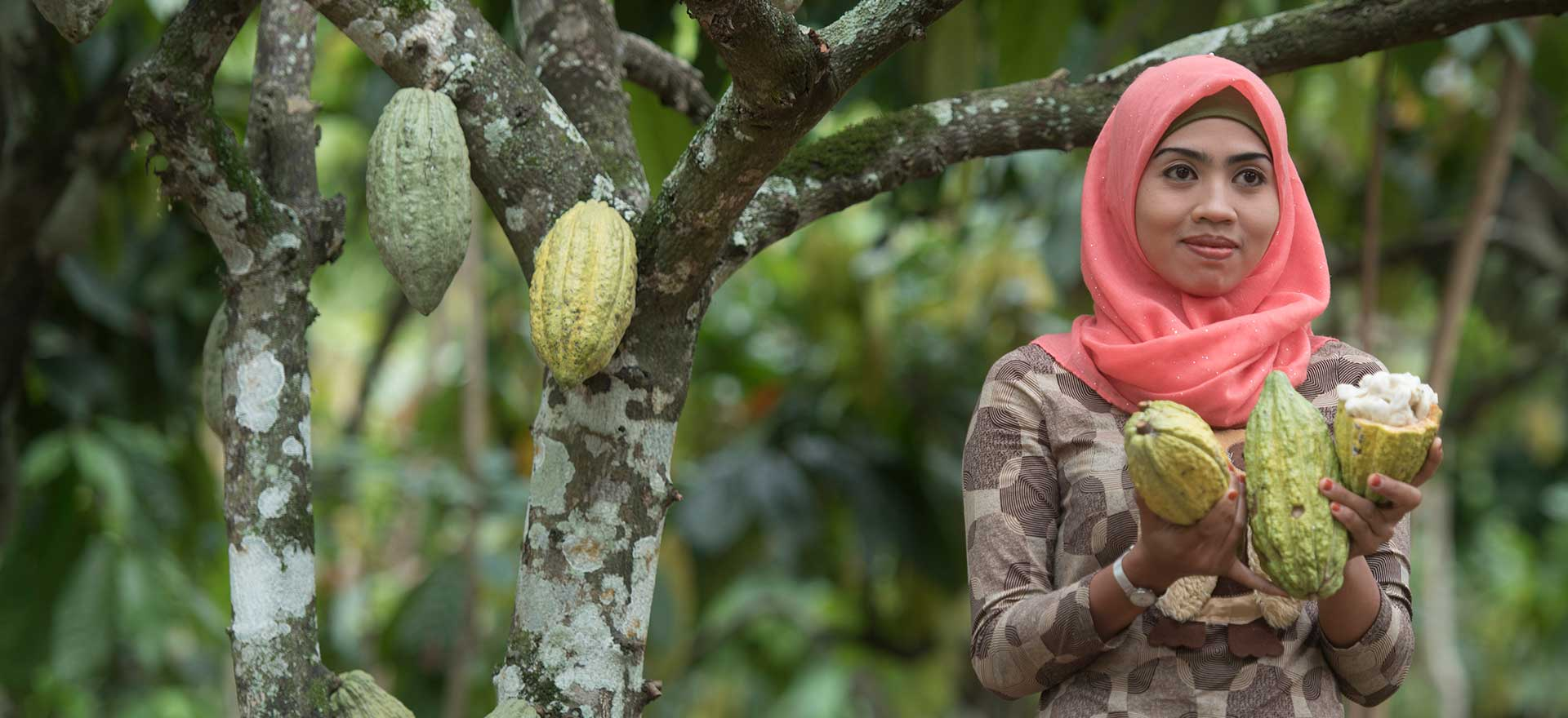 Indonesian woman smiling while standing next to a tree and holding fruits that have been picked from the tree.