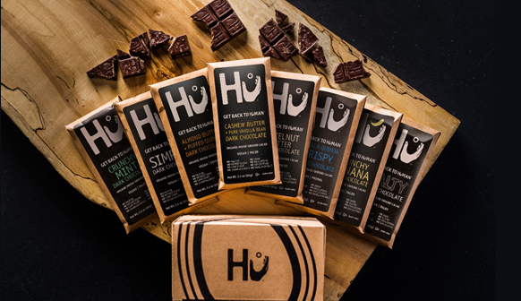 Overhead shot of 8 different flavours of Hu bars on a woodgrain table with various bits and pieces of individuals chocolate squares strewn about.