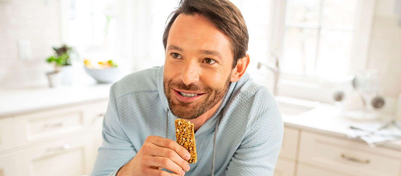 Man leaning over counter and smiling while eating a granola snack.