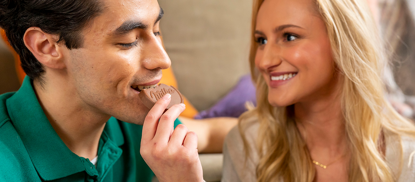 Woman and man smiling at each other while the man eats a Milka chocolate snack.