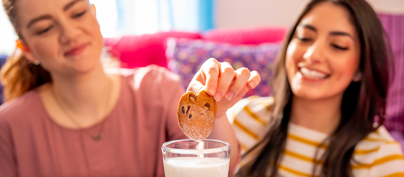 2 women smiling while one dunks a chocolate chip cookie into a glass of milk.