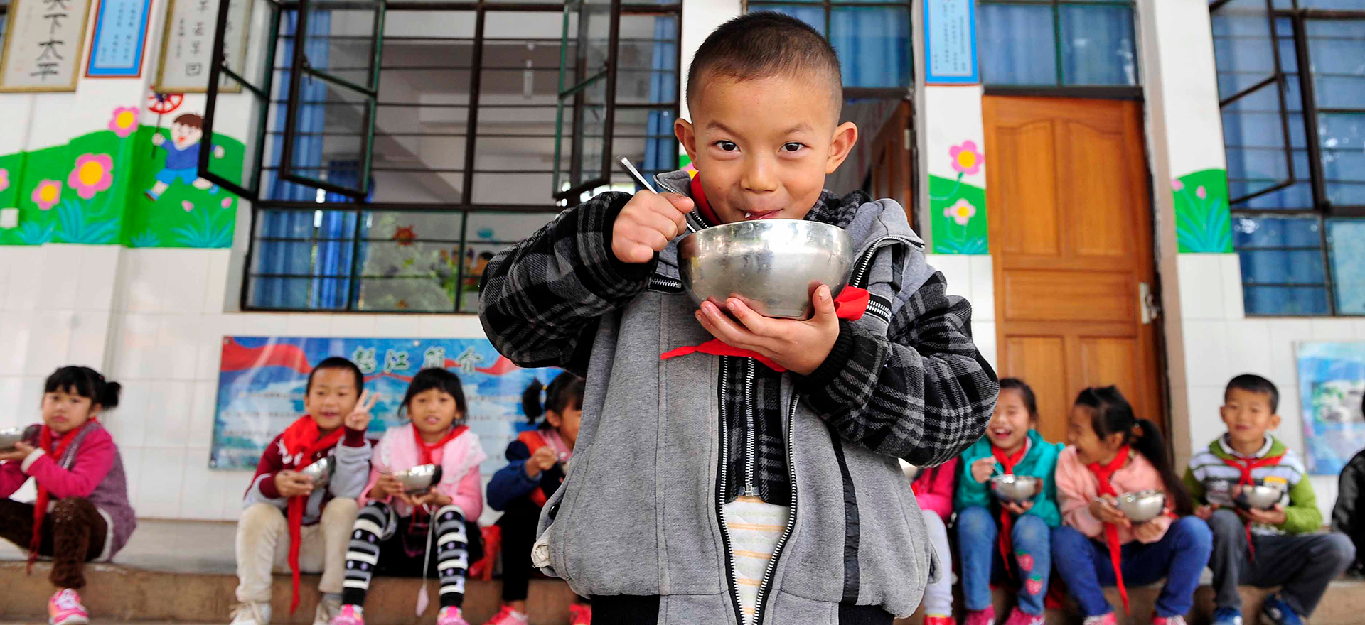 Asian children at a school sit down eating bowls of food while 1 stands in the foreground also eating from a bowl and smiling.